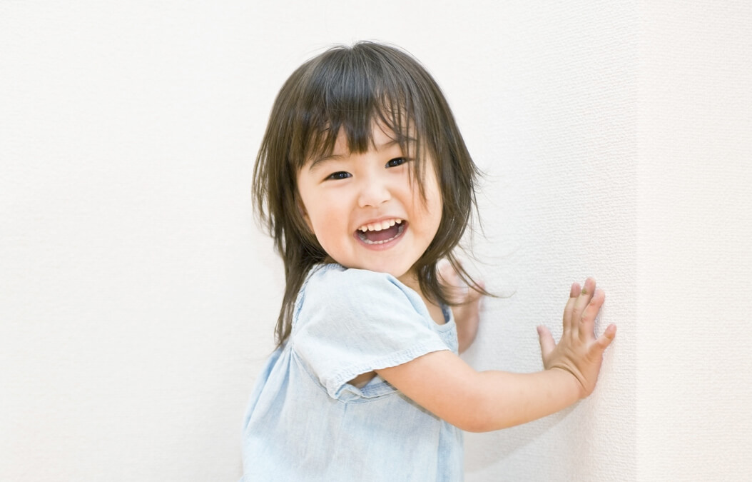 子供が壁に手のひらをつけて、こちらをみて笑っている様子